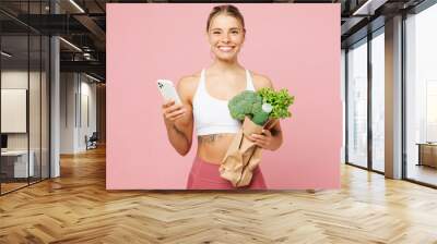 Young smiling woman wear white top casual clothes hold packet with green vegetables use mobile cell phone isolated on plain pink background Proper nutrition healthy fast food unhealthy choice concept Wall mural