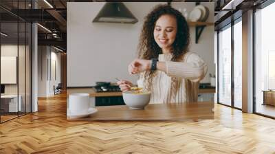 Young smiling woman wears casual clothes sweater eat breakfast muesli cereals with milk fruit in bowl look at smart watch sit at table in light kitchen at home alone. Lifestyle cooking food concept. Wall mural