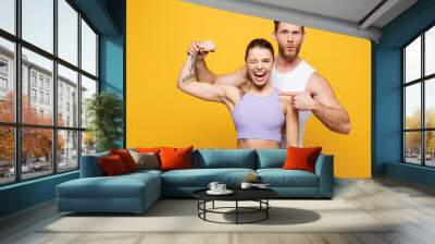 Young strong fitness trainer instructor sporty two man woman wear blue clothes spend time in gym point finger on biceps muscles on hand isolated on plain yellow background. Workout sport fit concept. Wall mural