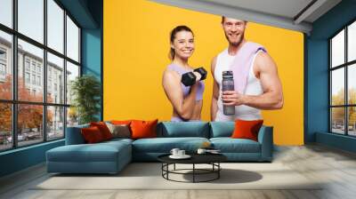 Young strong fitness trainer instructor sporty two man woman wearing blue clothes spend time in home gym hold in hand dumbbell bottle of water isolated on plain yellow background Workout sport concept Wall mural