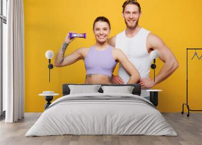 Young strong fitness trainer instructor sporty two man woman wears blue clothes spend time in home gym hold in hand protein energy bar isolated on plain yellow background. Workout sport fit concept. Wall mural