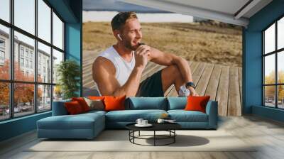 Young strong sporty athletic toned fit sportsman man he wears sports clothes eat protein bar drink water warm up train at sunrise sun dawn over sea beach outdoor on pier seaside in summer day morning. Wall mural