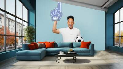 Young surpirsed man fan in t-shirt foam 1 fan glove finger up point overhead cheer up support football sport team hold soccer ball watch tv live stream isolated on plain pastel blue color background Wall mural