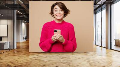 Young surprised cheerful smiling caucasian woman wearing pink sweater hold use mobile cell phone look camera isolated on plain pastel light beige background studio portrait. People lifestyle concept. Wall mural