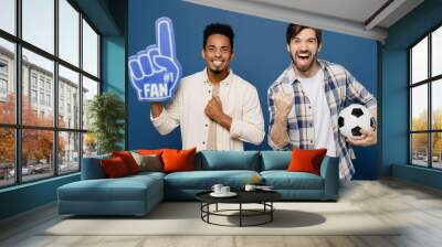 Young two fan friends cool men wear white shirt foam glove finger up support football sport team hold soccer ball watch tv live stream do winner gesture isolated plain dark royal navy blue background Wall mural