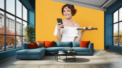 Young woman girl in casual clothes hold in hand makizushi sushi roll served on black plate japanese food using mobile cell phone isolated on yellow background studio portrait. People lifestyle concept Wall mural