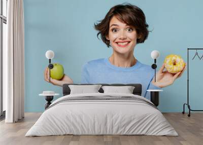 Young woman in casual sweater look camera hold donut dessert apple choose beatween healthy and junk food isolated on plain pastel light blue background studio portrait. People lifestyle food concept. Wall mural