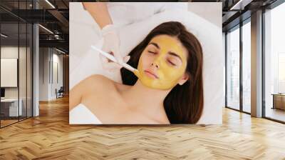 The cosmetologist applies a facial gold mask to the woman's face. Cosmetology and facial skin care in beauty salon. Gold mask. Cosmetic procedure.  Wall mural