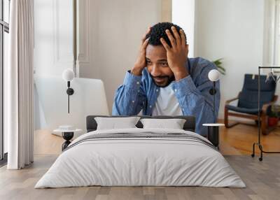 Frustrated young African entrepreneur with sad grimace in front of his laptop at home office, noticed a big mistake he did, holding his face with hands, wearing blue shirt Wall mural