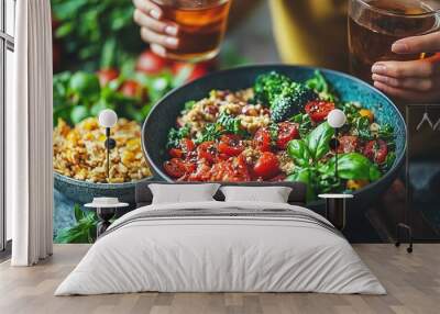 A colorful bowl filled with fresh vegetables and grains, as a person holds drinks nearby, celebrating a healthy meal Wall mural