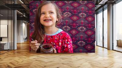 Cute happy young smiling girl eat homemade sweet chocolate paste spread from big glass jar with joy and pleasure Wall mural