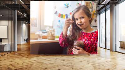 Cute happy young smiling girl eat homemade sweet chocolate paste spread from big glass jar with joy and pleasure Wall mural