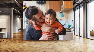 Hipster young father hug and kiss his baby infant boy holding on hands. At home on kitchen background. Lifestyle photo of family care and happiness concept. Middle class.  Wall mural