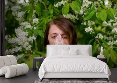 A beautiful girl stands by the blossoming bird cherry and sneezes. She suffers from an allergy to flowering in the spring. Wall mural