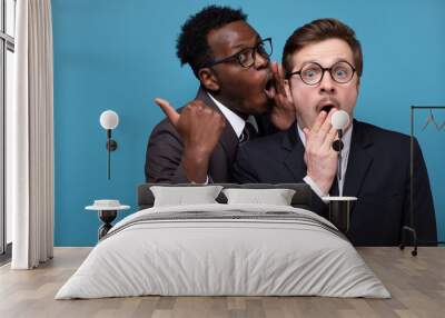 African man whisperingto his caucasian coworker ear telling him something secret. Studio shot on blue wall. Human emotions facial expressions Wall mural