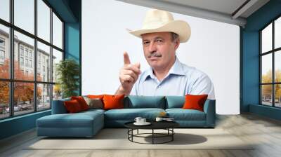 Serious mature hispanic man in cowboy hat showing index fingers up, giving advice or recommendation Wall mural