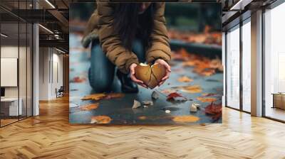 Woman kneels on wet autumn street holding crumbling heart in hands. Symbol of broken love and separation from partner. Heartbreak and sorrowful emotions in human relationships. Wall mural