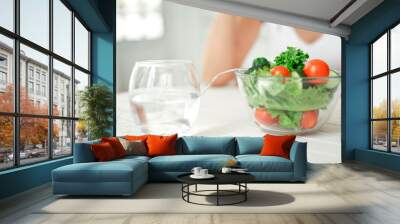 Girl is sitting at the table and going to eat some healthy meal. Bowl of salad and glass of water on the white table. She is happy smiling in white bright room. The picture is cropped to highlight Wall mural