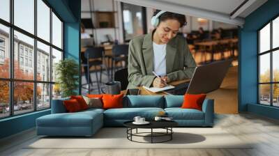 A woman in a hat with headphones sits at a laptop on a wooden desk in nice modern coworking office Wall mural