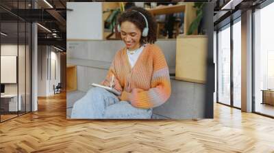 a woman wearing headphones is sitting on the floor writing in a notebook Wall mural