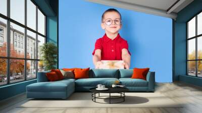 Portrait of a happy cute little boy wearing eyeglasses, holding a gift box and looking into a camera isolated on a blue background. Wall mural