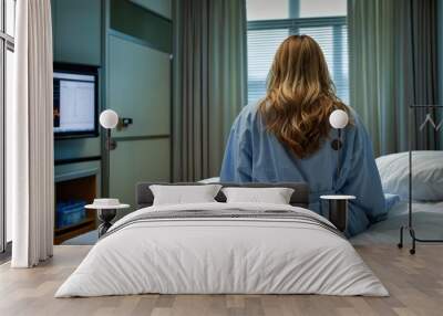 Back view of a woman in a hospital gown sitting on a bed, looking out a window, creating a calm yet reflective atmosphere in a medical room.
 Wall mural