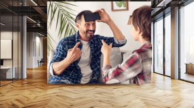 Father and little son at home sitting on sofa playing heads up with smartphone guessing word cheerful Wall mural