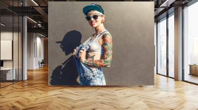 Freestyle. Alternative girl in cap ans sunglasses standing isolated on grey on the city street playing with short overall smiling Wall mural