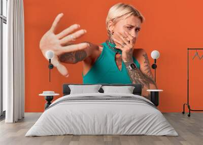 Freestyle. Young tattoed woman standing isolated on orange hand forward protective covering mouth scared close-up Wall mural