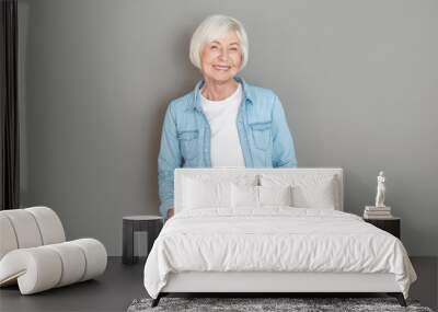 Senior woman in jeans jacket studio isolated on grey wall hands in pockets joyful Wall mural