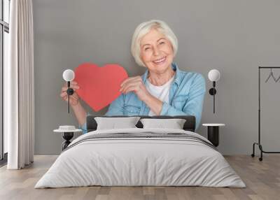 Senior woman in jeans jacket studio isolated on grey wall saint valentine's day holding red heart happy Wall mural