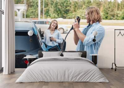 Transportation. Young couple traveling by car having stop at charging station boyfriend playing with plug looking at girlfriend drinking hot coffee laughing cheerful Wall mural