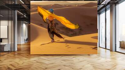 Beautiful young slim woman in a multi-colored dress with a yellow scarf, in a turban and sunglasses poses at dawn in the Sahara desert. Morocco Fashion. Wall mural