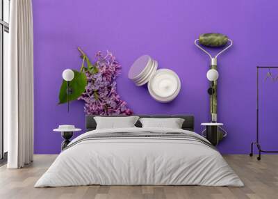 Jade facial roller, cream jar and lilac flower on purple background. Natural cosmetics, skin care concept. Top view, flat lay Wall mural