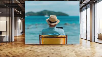 A man in a straw hat sits on a chair facing the ocean, working on a laptop while enjoying a tropical beach with an island view in the background. Wall mural