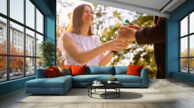 Joyful young woman volunteer in white blouse looking at the homeless man and giving him a plastic bottle of water to drink. Wall mural