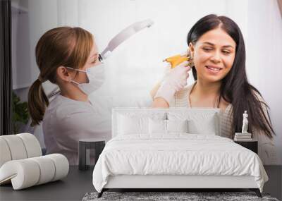 Specialist in medical mask and gloves pierces the client's ears with a piercing gun in the cosmetologist cabinet. Wall mural