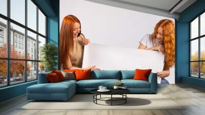 Two red-haired girls are holding a large white poster that might contain your ad. Photo on white background Wall mural