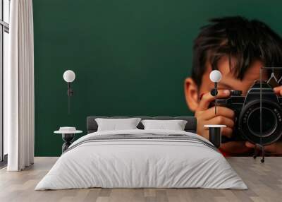 A young boy holding a camera, looking through the viewfinder, in front of a green background. Wall mural