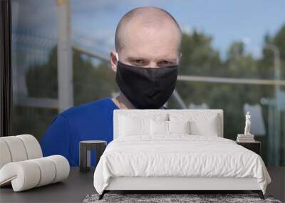 Male in a black medical mask looks into the camera lens, in the background you can see an iron fence. Wall mural