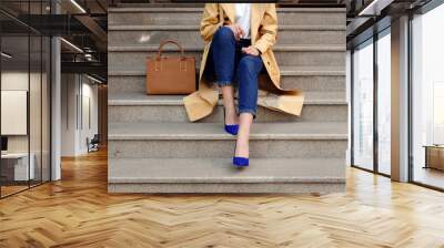 Street fashion outfit, model in blue jeans coat and brown handbag in high heel shoes sitting on the beige stair, outdoor Wall mural