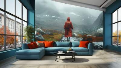 A female hiker in a large long red raincoat is walking through a wide alpine valley covered in boulders in mountains, it's raining heavily and she is pushing against the rain and strong winds. Wall mural