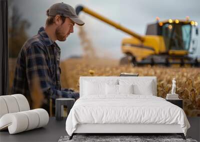 A young farmer in a field with his laptop in the foreground, looking at an app on it. behind him is a modern yellow harvester is working in the background. Generative AI. Wall mural