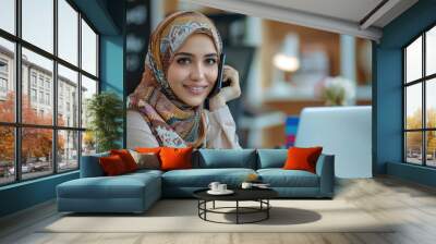 Muslim woman in hijab is sitting at her desk and talking on the phone, smiling as she looks up from working with her laptop. Generative AI. Wall mural