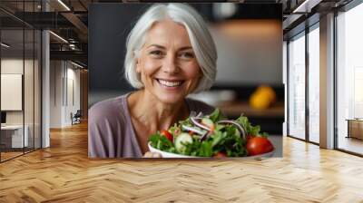 Adult vegetarian woman in casual attire smiling holding a healthy vegetable salad bowl on blurred kitchen background Wall mural