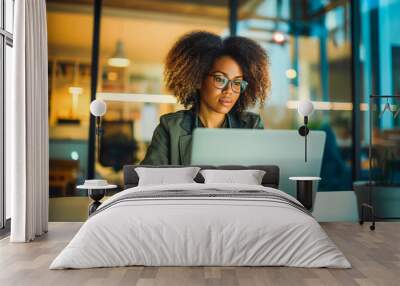 A confident young Afro-American woman typing on her laptop in a modern office flooded with natural light, her determined expression illuminated by the glow of her computer screen. Wall mural