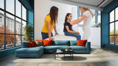 A young girl patient sits on an examination table in a medical office, while a healthcare professional doctor checks her heartbeat with a stethoscope. A woman stands nearby, observing. The room is bri Wall mural