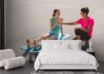 A young woman sitting on a yoga mat, smiling and holding a water bottle, while an older woman kneels beside her, offering encouragement. They are on an outdoor sports court. Wall mural