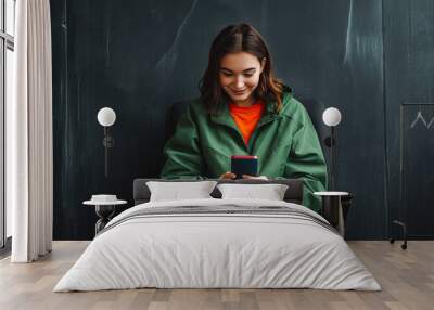 Happy relaxed young woman wearing green jacket sitting on modern chair using phone against dark background. Wall mural