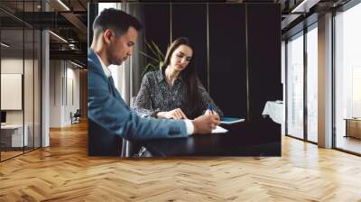 Two business people in business clothes are sitting in an office looking at a contract. One person is giving advice to the other. After signing the contract, they shake hands. Together they look out o Wall mural